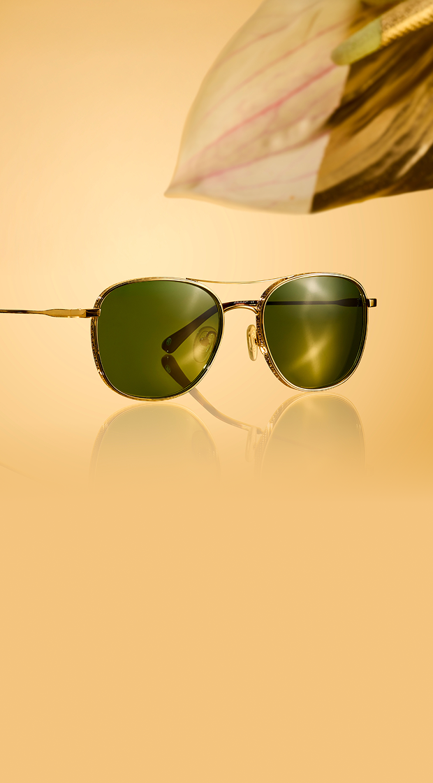 Gold rim aviator sunglasses with green tinted lenses on a reflective surface against a warm golden background.