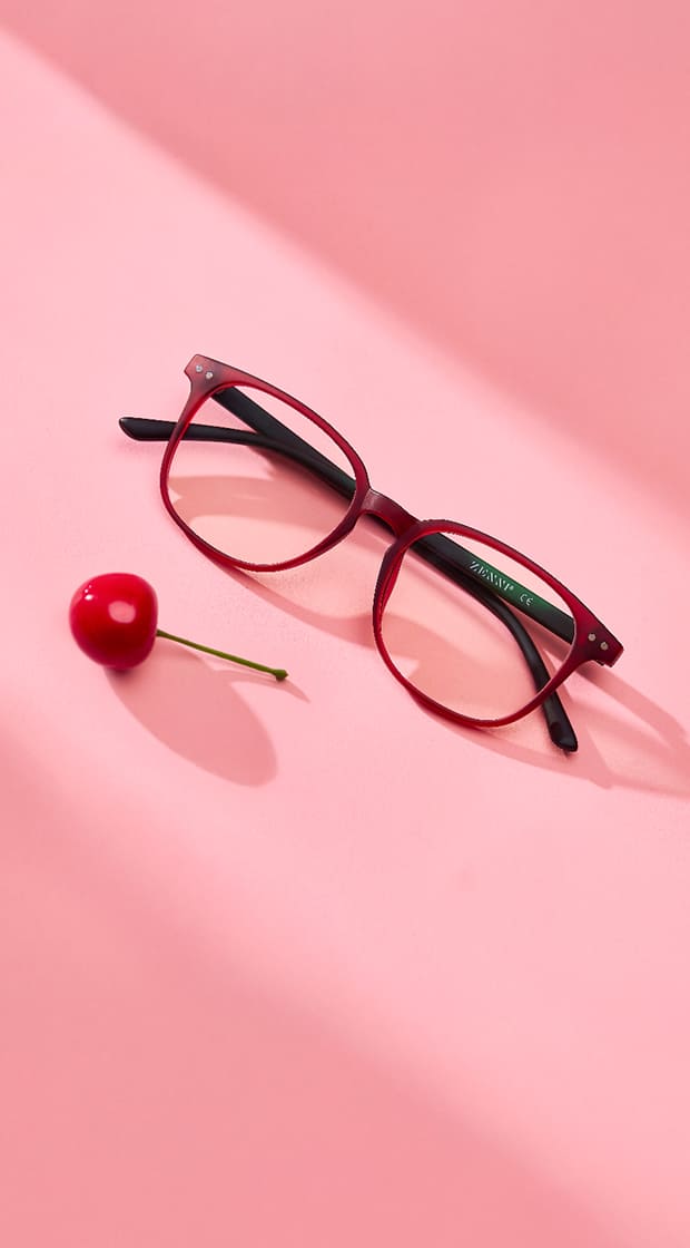 A pair of red eyeglasses with black arms lies on a pink surface surrounded by three cherries.