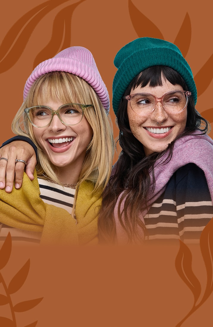 A blond woman wearing clear green glasses and a pink knit hat being hugged by a brunette woman wearing clear pink glasses and a green knit, on a brown background with a foliage motif.