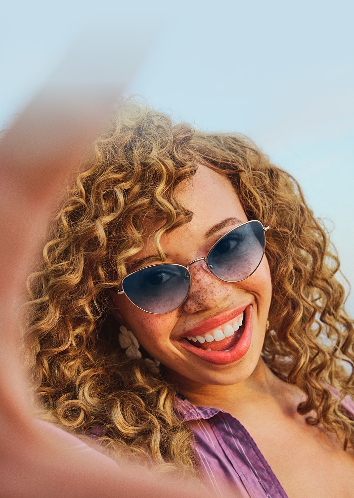A woman with freckles and curly dark blond hair wearing wire frame cat-eye sunglasses with a blue gradient tint.