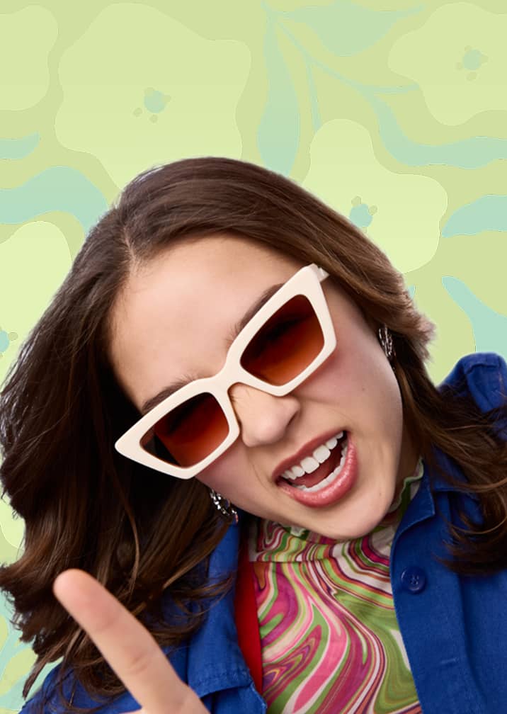 A woman with wavy brown hair and an excited expression, wearing white cat-eye sunglasses with red tint lenses, on a green background with an abstract flower motif.
