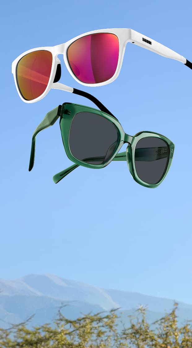 Two pairs of sunglasses floating in the air against a clear blue sky, including white sport sunglasses with reflective red lenses and translucent green square sunglasses, with a blurry mountain landscape below.