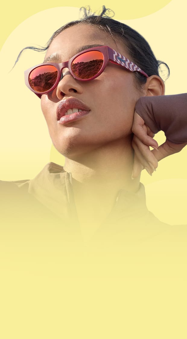 Close-up of a woman looking away while wearing matte red cat-eye sunglasses with reflective orange lenses, set against a light yellow background with abstract wavy patterns.