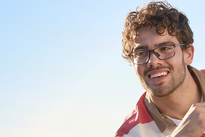 A man with curly hair wearing rectangle sports Zenni eyeglasses with clear lenses. Blue sky background.