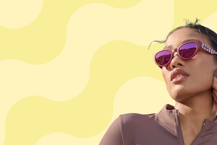 A woman wearing sporty pink Zenni sunglasses with patterned temples and pink mirror lenses on a yellow wavy stripe background.