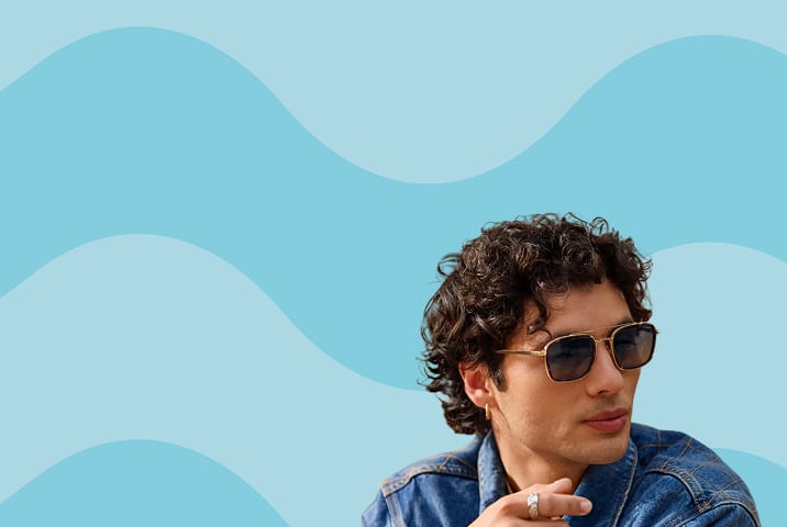 A man with brown curly hair wearing gold aviator Zenni sunglasses. Blue wavy stripe background.