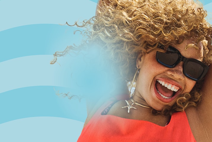 A woman with curly light brown hair smiling broadly while wearing square black Zenni sunglasses with dark lenses. Blue wavy stripe background.