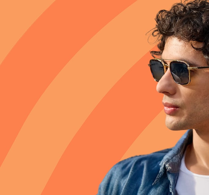 Profile view of a man with wavy dark hair wearing a denim jacket and square wireframe sunglasses, set against a vibrant orange background with abstract curved stripes.