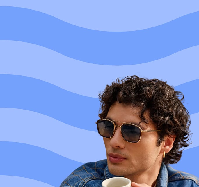 Close-up of a man with wavy dark hair wearing a denim jacket and gold-trimmed navigator sunglasses while holding a coffee cup, set against a light blue background with dark blue waves.