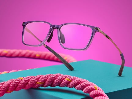 A pair of translucent purple eyeglasses rests on a teal rectangular block along with a pink braided ropes with yellow accents, against a vibrant pink background.