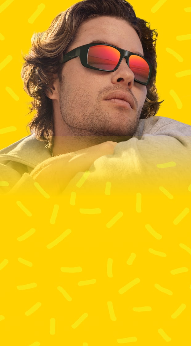 Chase Stokes wearing black sport-style sunglasses with reflective red-orange lenses, leaning forward with arms crossed against a bold yellow background with playful squiggle patterns.