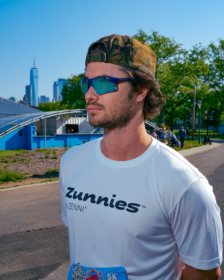 Chase Stokes wearing mirrored Zunnies sunglasses and Zunnies shirt running outdoors with city skyline in the background.
