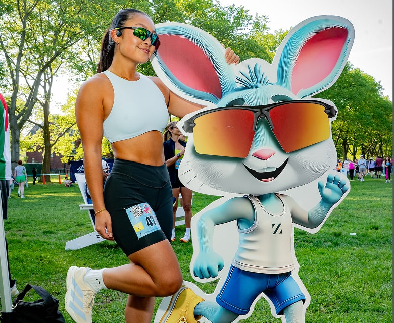 Woman in athletic wear posing next to a large Ziggy mascot cutout wearing Zunnies sunglasses.