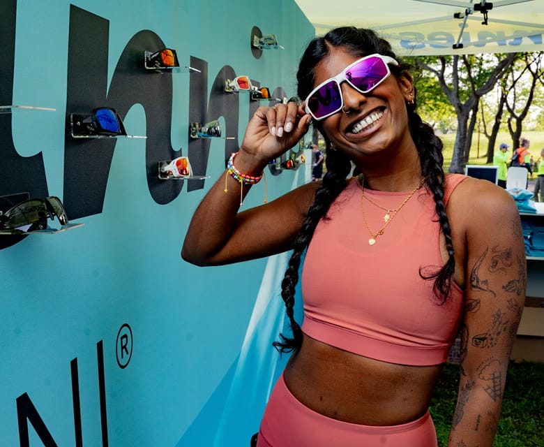 Woman in coral activewear smiling and wearing Zunnies sunglasses in front of an event display wall.