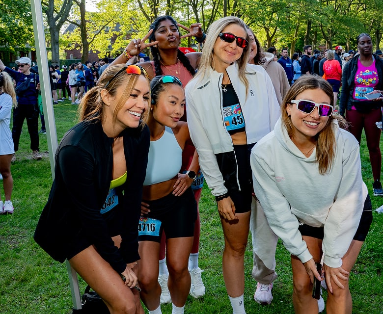 Group of women in athletic wear laughing and posing for a photo at a sunny outdoor race event, wearing Zunnies running sunglasses.