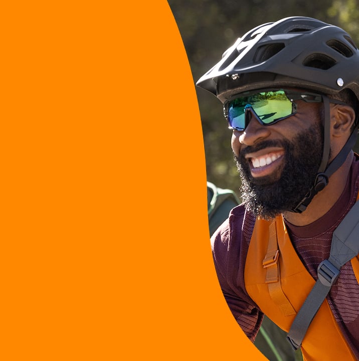 A smiling man wearing reflective sunglasses and a bike helmet is geared up with a backpack, ready for a cycling adventure.