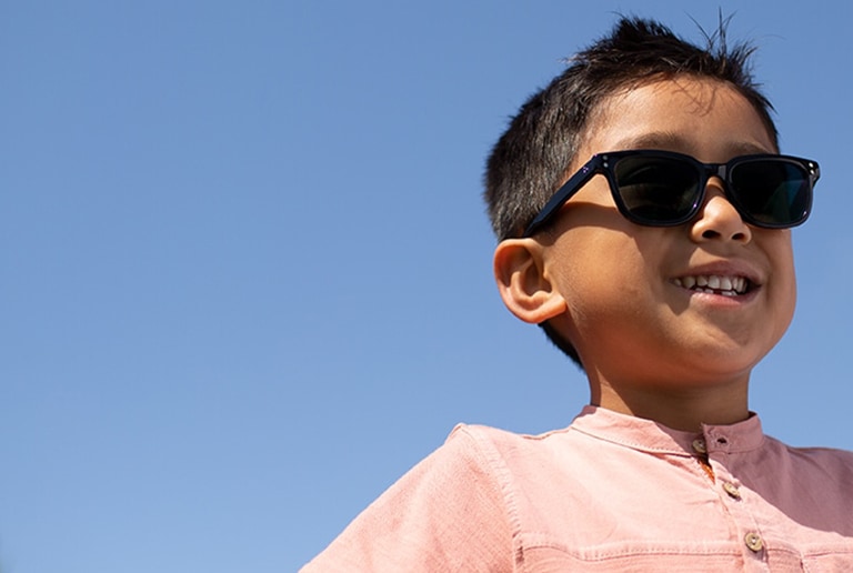 Young boy with short brown hair standing outside wearing Wayfarer-style black acetate sunglasses #4433121 with photochromic lenses and a pink short-sleeve pocket tee.