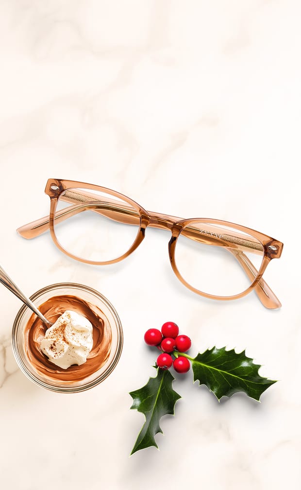 A pair of light brown eyeglasses beside a creamy coffee drink topped with whipped cream and festive holly berries.