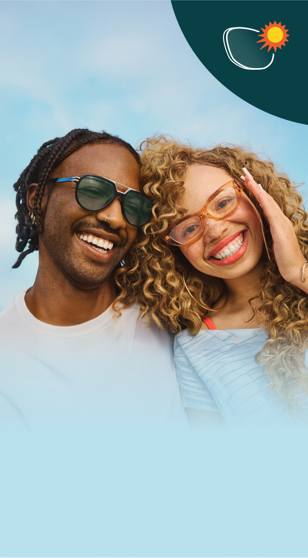 Two people smiling outdoors under a bright sky, both wearing glasses. The man’s lenses are tinted dark from sunlight while the woman’s lenses are clear, showing how photochromic lenses adapt to light.