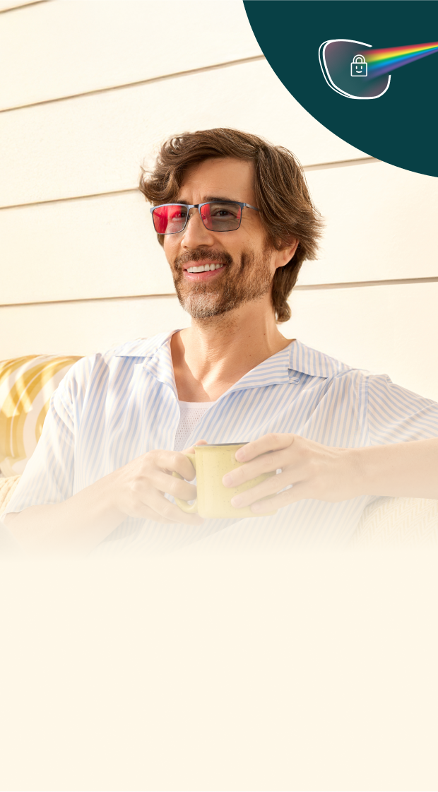 A man with medium-length brown hair and beard is wearing stylish pink-tinted glasses while sitting on a couch in a sunlit room. He is smiling and holding a yellow pillow. In the upper-left corner, there is a dark green semi-circle with a white eyeglass icon and a rainbow light beam symbol with a lock, suggesting light filtering or eye protection technology.