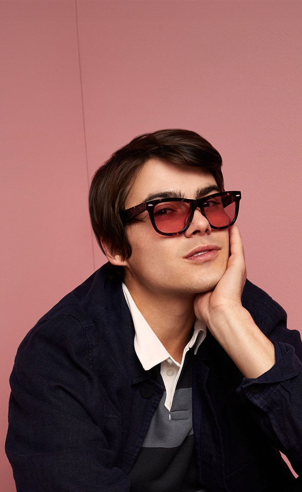 A thoughtful man in a dark jacket and white collared shirt wearing oversized Zenni FL-41 rose-tinted glasses, seated against a pink wall.