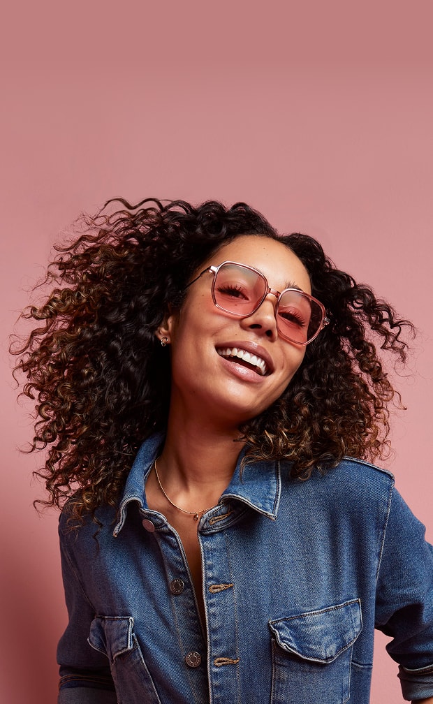 Woman with curly hair smiling, wearing a denim jumpsuit and Zenni FL-41 rose-tinted glasses, against a pink background.