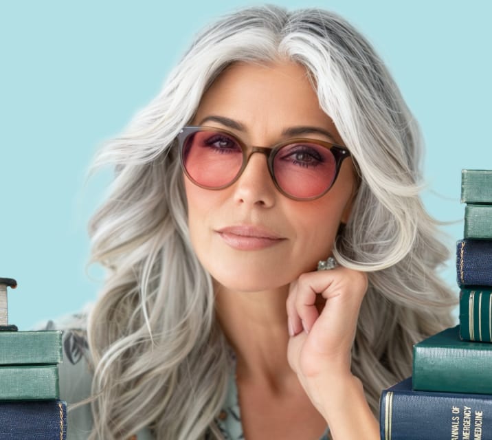 A woman with long gray hair wears circular readers glasses and smiles softly at the camera, her chin resting on her hand. The background is light blue, and green and blue books are stacked on both sides of her.