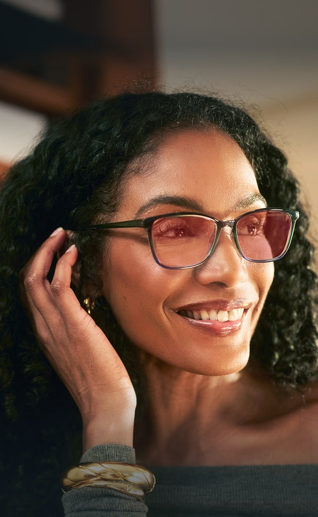 A woman with curly black hair smiles warmly while holding one hand near her face. She is wearing rectangular glasses with light pink-tinted lenses and a gray off-shoulder top, accessorized with gold bracelets.