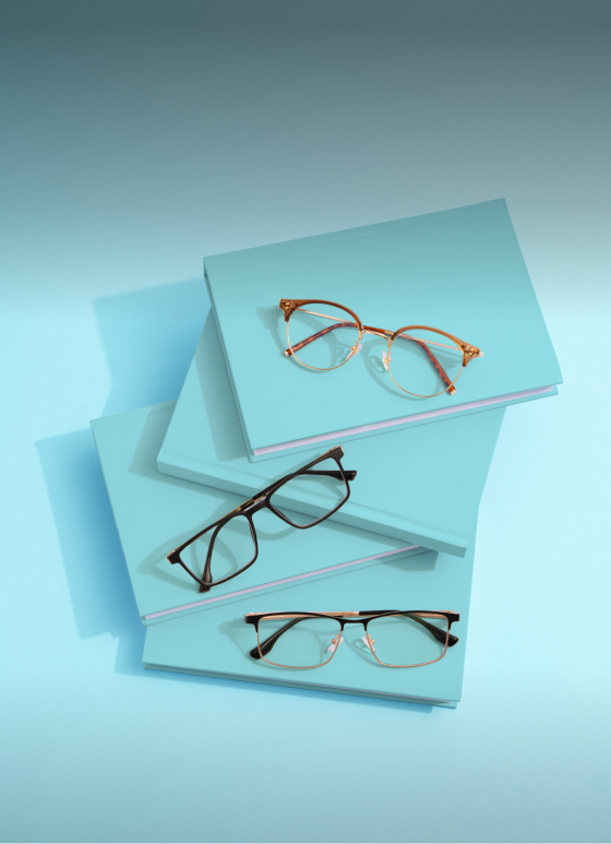 Three pairs of lightweight reading glasses in geometric shapes arranged on a light blue surface. Above, text reads "Lightweight Readers" and "For ultimate comfort." At the bottom, a white rectangular button says "Shop now." The background includes a rolled sheet of blue paper.