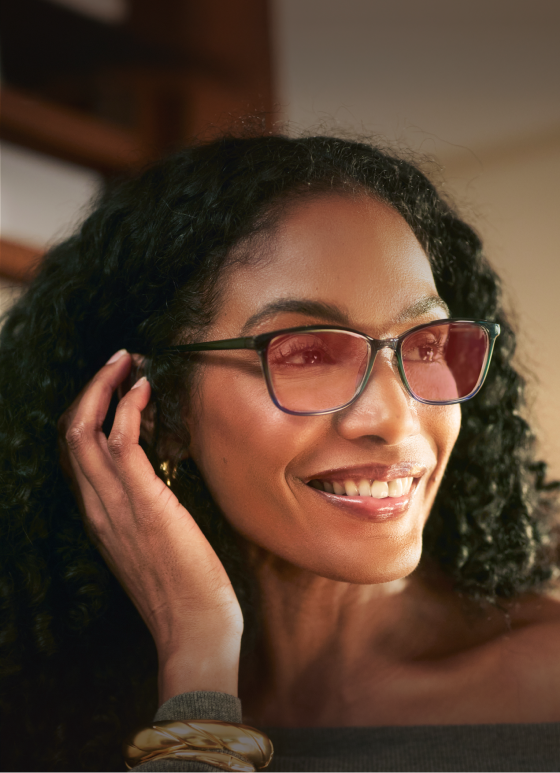 A woman with curly black hair smiles warmly while holding one hand near her face. She is wearing rectangular glasses with light pink-tinted lenses and a gray off-shoulder top, accessorized with gold bracelets.