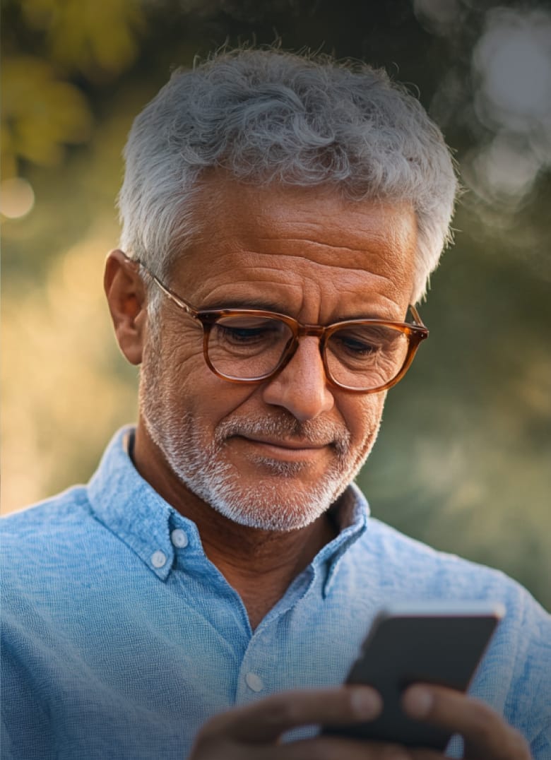 An older man with short, curly gray hair and a trimmed beard looks at a smartphone while wearing round brown glasses. He is outdoors with blurred greenery in the background.