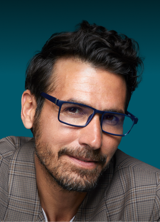 A man with dark hair, wearing blue light protection readers glasses in a rectangular shape and a checked blazer, smiles at the camera. Text above reads "Blue Light Protection" and "Add Blokz® to your readers." At the bottom, a white rectangular button says "Shop now." The background is dark teal.