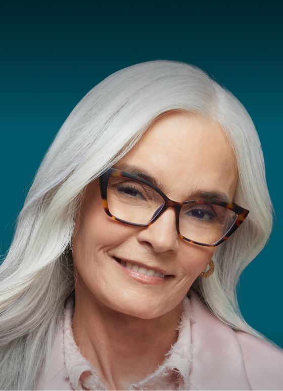 A woman with long white hair wearing cat-eye readers glasses smiles softly at the camera. Text above her reads "Designer Readers" and "Deluxe, starting at $35." A white rectangular button with "Shop now" is displayed at the bottom. The background is a gradient of dark teal and light blue.