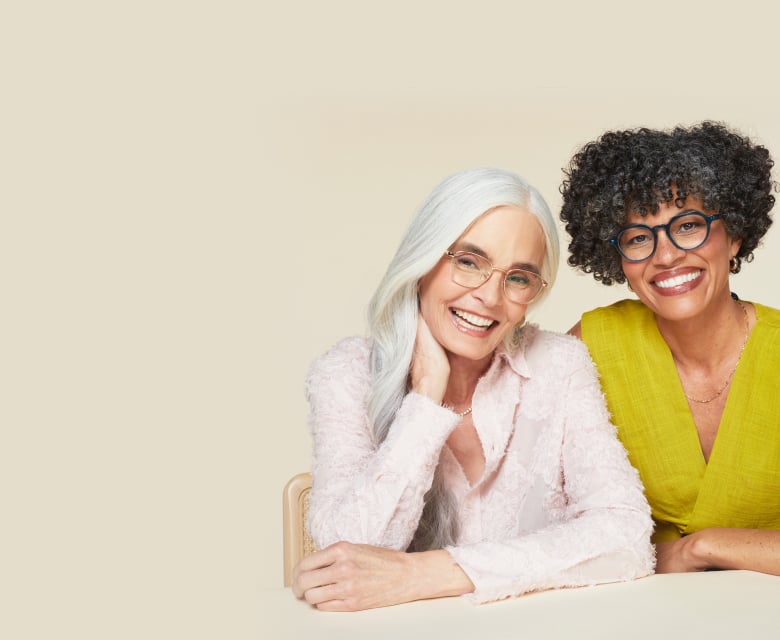 Two women sit together, smiling at the camera. One has long white hair and is wearing lightweight readers glasses, while the other has short curly hair and dark-framed readers glasses. Text beside them reads "Customizable Readers" and a white rectangular button at the bottom displays "Shop now." The background is beige.