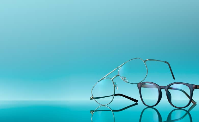 A pair of lightweight, metal-framed glasses and a pair of sleek, round, acetate-framed glasses are displayed on a reflective turquoise surface against a gradient blue background.